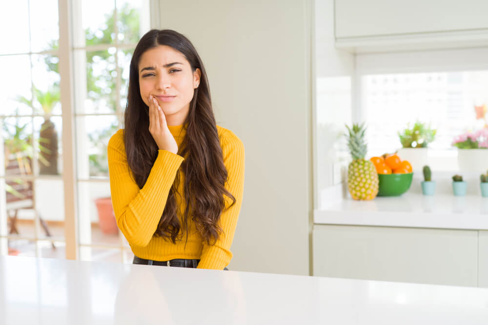 Woman with yellow sweater holding her right hand to her cheek in pain because of a tooth ache