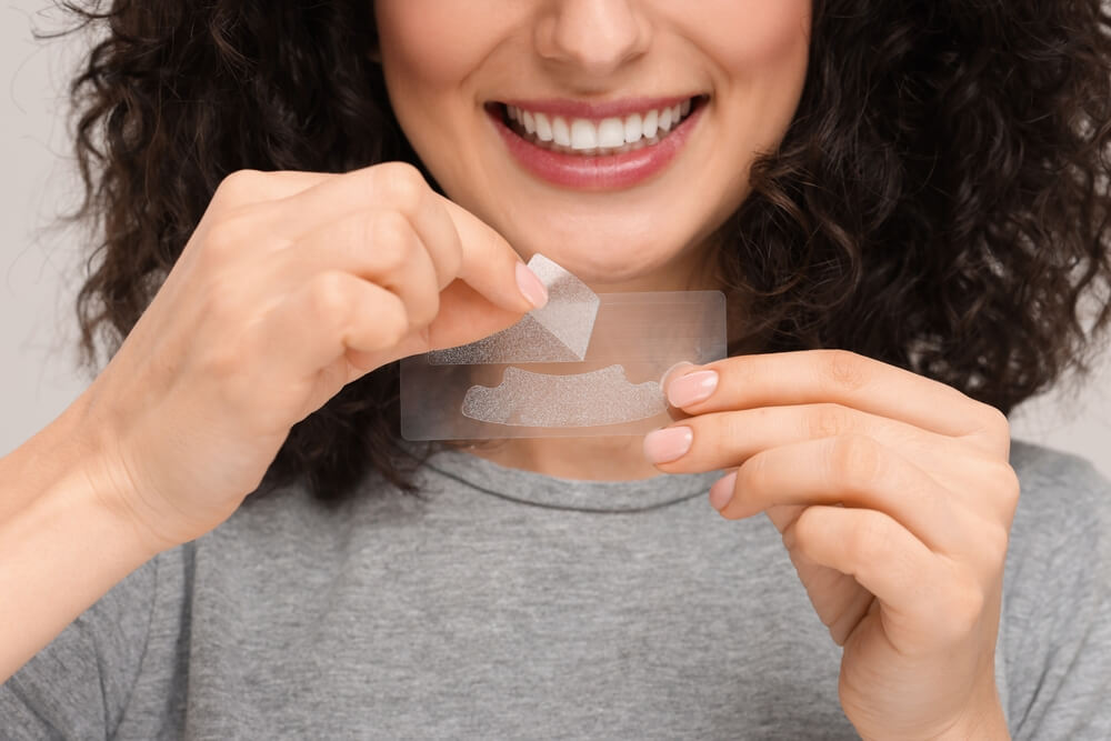 Woman peeling off a teeth whitening strip from a sheet, preparing for a cosmetic dental treatment to enhance her smile.