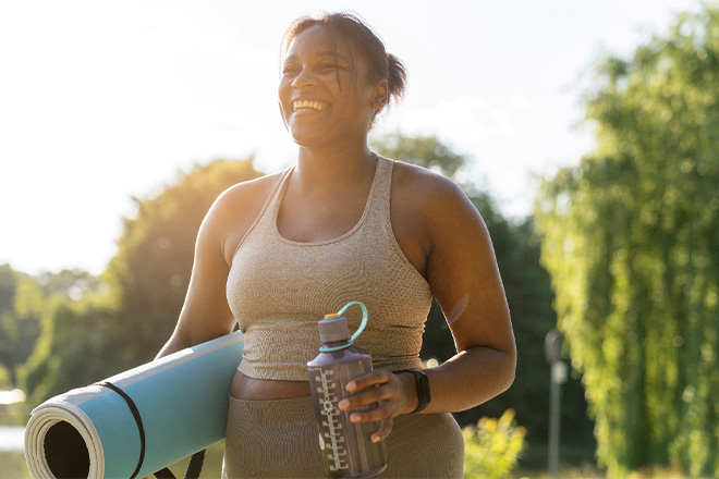 Woman smiling while holding workout mat and water bottle.